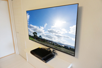 「白」の多い明るい雰囲気のリビングにブラビア・55インチ「KJ-55X8500E」の壁掛けとブルーレイレコーダー用の専用シェルフを同時に施工させて頂きました。
オーナー様は壁掛けテレビへのキッカケに、「省スペースさ」を重視されたと仰っていました。
生活動線上で、テレビ台を設置してしまうとどうしても狭さを感じてしまう点と、掃除のしやすさも意識されてご依頼されたとお伺い致しました。
事前のコンサルでは、テレビ台をどうしても置きたくないので、周辺機器の置き場所に悩まれていましたが、専用のシェルフをご提案させて頂く事で、解決致しました。
リビングへの入口から正面にテレビの側面が見えてしまう事もあり、テレビに合わせて金具を薄型タイプに選定し、テレビの壁からの出幅も大きく抑えて仕上げさせて頂きました。
シェルフ出幅も25cmとテレビ台と比較してもスッキリとした仕上がりに大きく貢献できています。
施工後には求められていた省スペースさを強く実感して頂く事が出来まして、見た目のカッコよさにもご夫婦から喜びの声も頂くことができ、大変嬉しく思います。